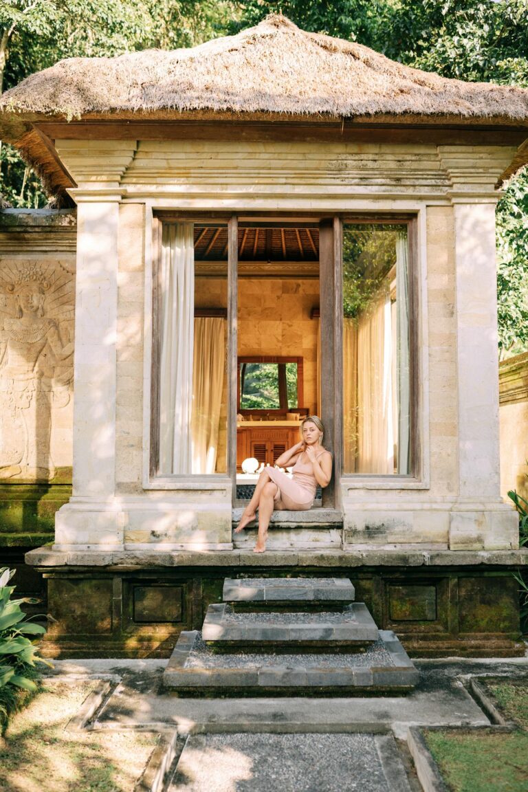 A woman sits elegantly on stone steps of a tranquil stone house with lush greenery.
